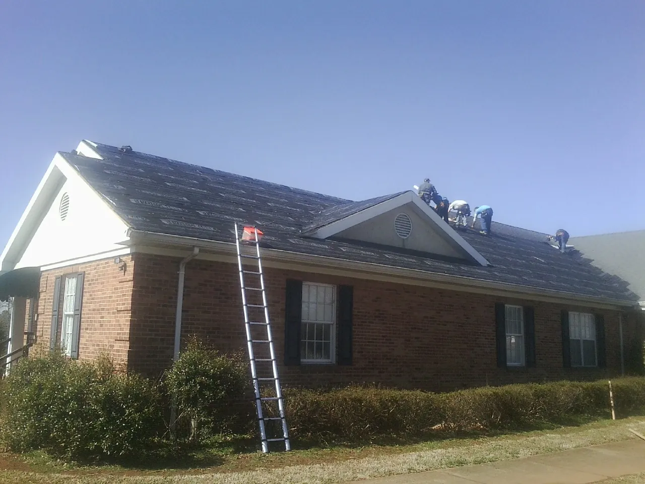 Crew tearing shingles off a brick ranch roof with a ladder set — Emergency Roof Repair work by our crew in Lake City
